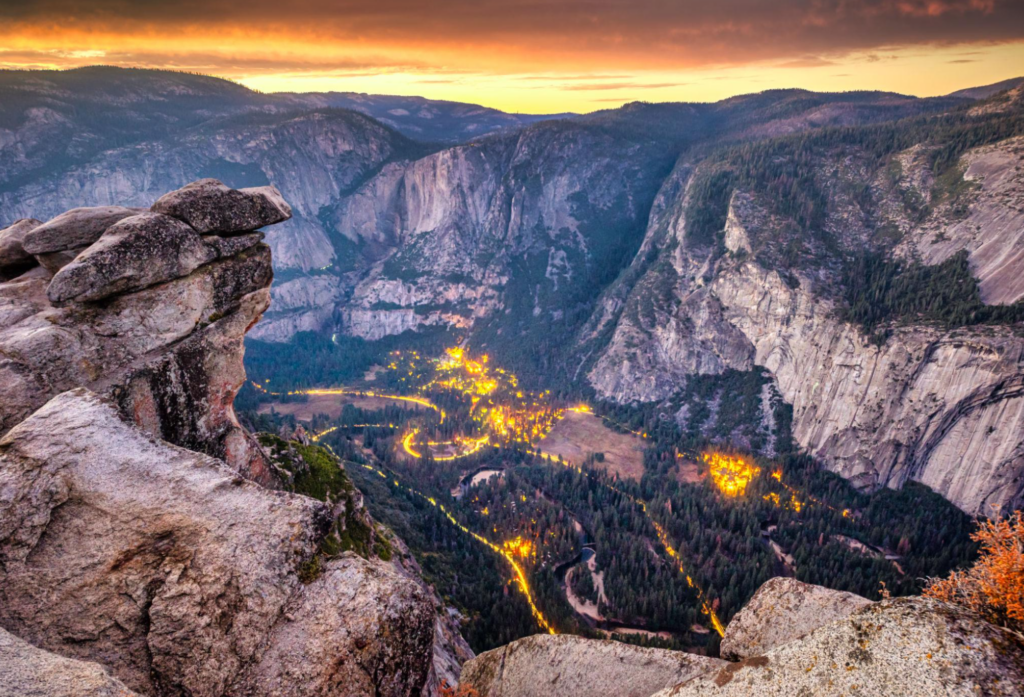 Vista elevada do Yosemite ao entardecer com luzes brilhando no vale abaixo e formações rochosas ao redor.