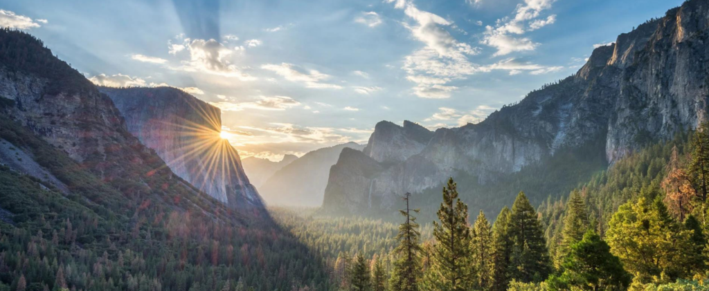Raio de sol atravessando as montanhas do Yosemite com floresta verde em primeiro plano.