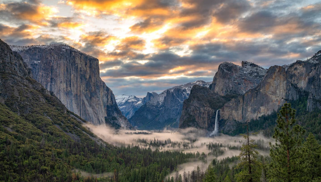 Vista panorâmica do Tunnel View no Yosemite ao amanhecer, com nuvens dramáticas, montanhas de granito e névoa sobre a floresta.